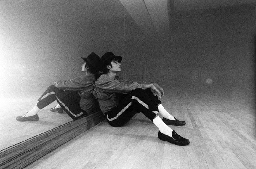 Michael Jackson sitzt nachdenklich am Boden eines leeren Tanzstudios, den Rücken an einen Spiegel gelehnt.